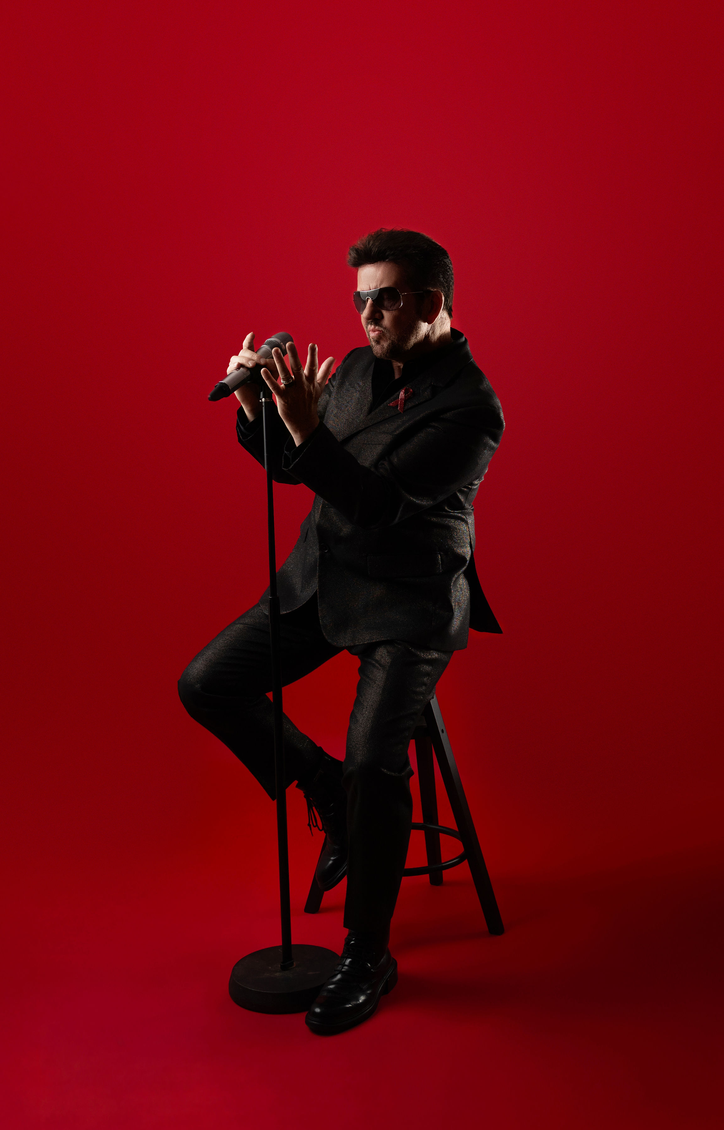 Steve Mitchell sitting on stool with microphone against red background