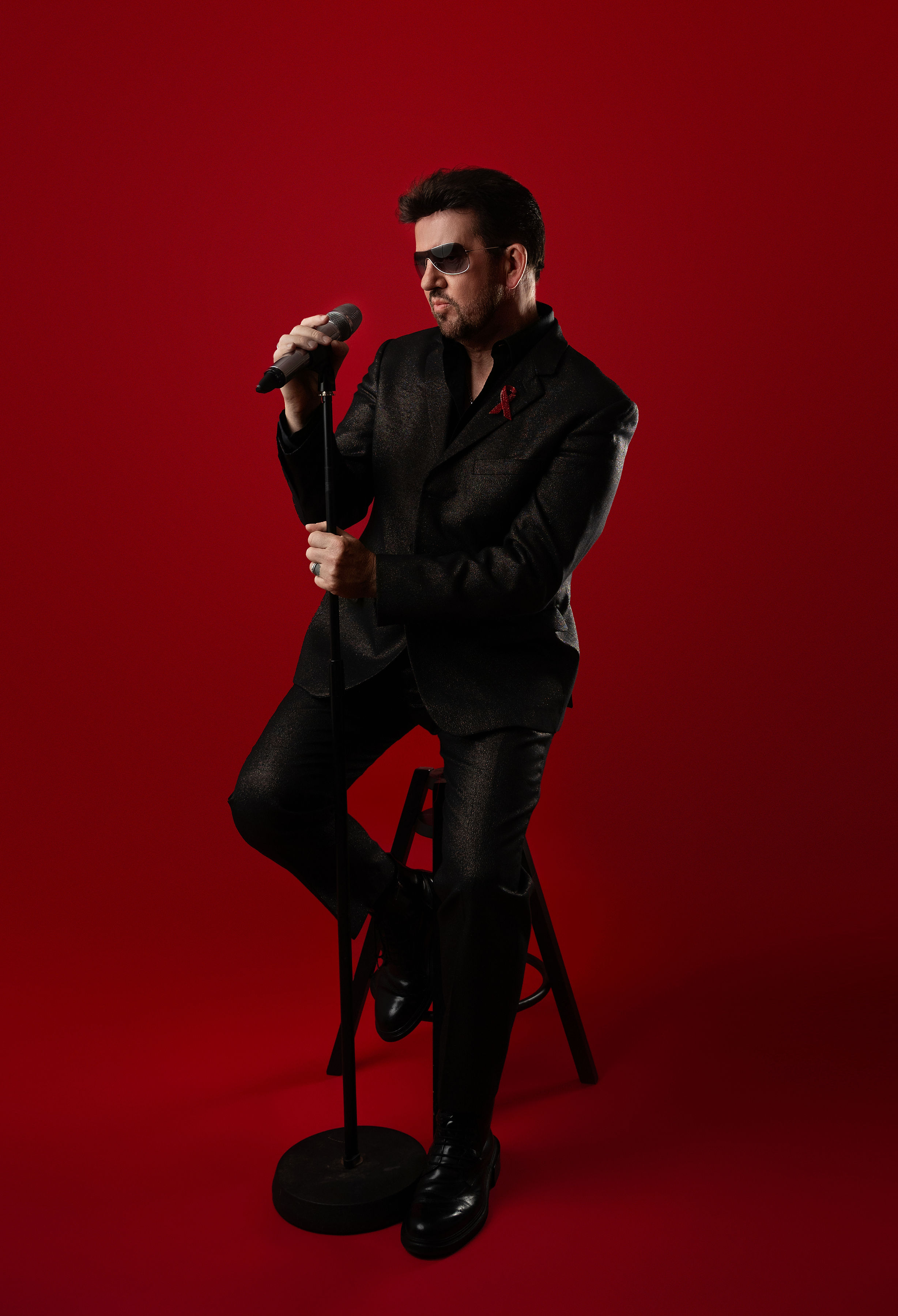 Steve Mitchell sitting on stool looking at microphone against red background